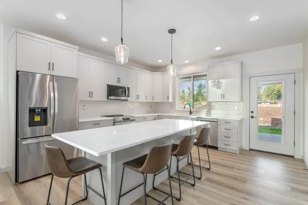 Kitchen in plan 280263JWD with white shaker cabinetry, a long quartz island with four stools, stainless appliances and a back door to the patio